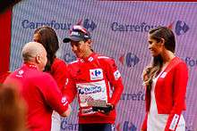 A photograph of Esteban Chaves, wearing the race leader's red jersey, shaking hands with an official, with podium girls either side.