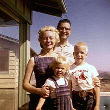 A man, woman, and two children smiling outside of a house