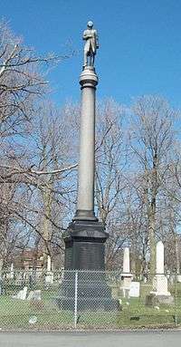 A tall stone monument behind a chainlink fence in a cemetery. A tall gray round column with a statue of a man atop sits on a darker pedestal