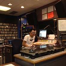 A DJ wearing sunglasses speaks into a microphone, surrounded by audio equipment, a computer, LP players, a darkened glass window, and a soundboard.
