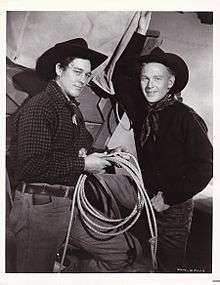 Black and white photograph showing two men standing. They are dressed in cowboy garb: jeans, kerchiefs around their necks, and cowboy hats. The man on the left is holding a lasso near his waist. A section of what appears to be a covered wagon is visible behind them.