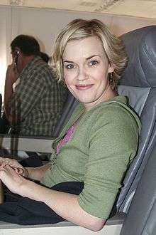 A blonde-haired young woman sitting in an airplane seat in a green top smiling at the camera.
