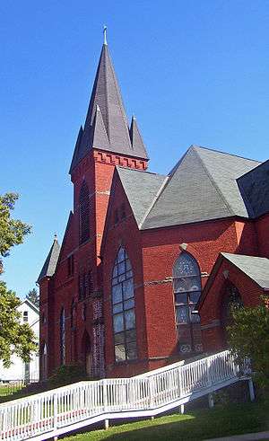 A red brick church with several peaked black towers and a white wooden wheelchair ramp in front