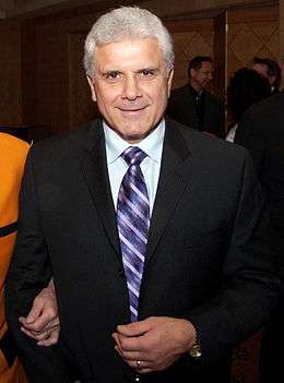 An Italian-Canadian man in his sixties with grey hair, wearing a tuxedo with a light blue shirt and purple tie, facing the camera.