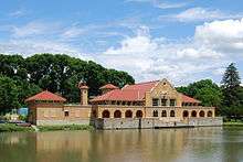 A yellow brick building, seen from across a lake, with red tiled roofs supported by brackets. It has a wide tower at the left end, arcade and ornate parapet in the center, and taller towers in the rear. There are taller trees behind it.