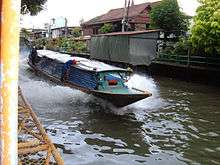 A boat, about ten metres long, travelling along a canal, the dark water breaking up in foam as it passes