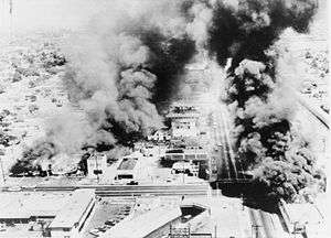 Several buildings burning in an aerial black and white photograph.