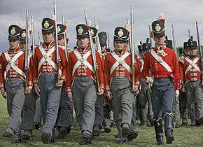 Photo shows marching men carrying muskets. They are dressed in red coats with white cross belts, gray trousers, and black shakos with brass frontplates.