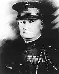 A black and white image of Wendell C. Neville, a white male in his Marine Corps dress blue uniform. He is wearing a hat and his French Fourragère can be seen, in addition to several ribbons.