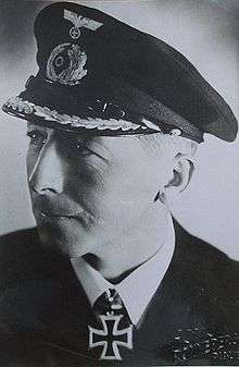 The head and shoulders of a man, shown in semi-profile. He wears a peaked cap, white shirt and a black jacket, and a military decoration in shape of an Iron Cross at his neck.