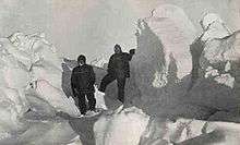  Two men in heavy clothing stand surrounded by mounds of ice which extend well above the height of their heads