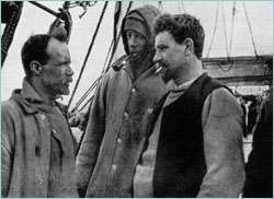  Three men in well-worn clothing stand on the deck of a ship. Man on right is thickset, bearded, with cigarette in mouth. Man in centre is hooded; man on left, also bearded and with receding hair, is shown right profile.