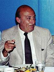 An bald elderly man in a suit and tie, seated before a meal, raising his right hand slightly and look to the right of the picture.