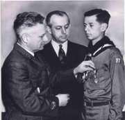Two men pin an Eagle Scout medal onto a young man wearing a 1940s era Boy Scout uniform