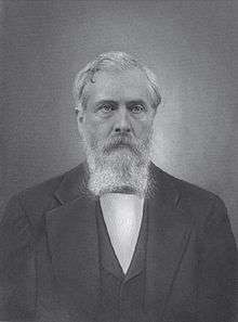 A man with white hair and a white beard and mustache wearing a white shirt, black jacket and black vest