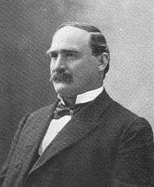 A man with receding, black hair and a thick, black mustache wearing a black jacket and tie and white shirt