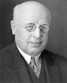 A photographic portrait of a balding white man with small round glasses