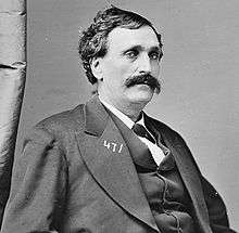 A man with black, curly hair and a mustache wearing a black jacket, vest, and tie and white shirt