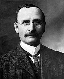 A photographic head and shoulders portrait of a moustached man wearing a three piece suit and round glasses