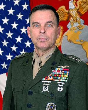 A color image of William Nyland, a white male in his Marine Corps Service A uniform. He is not wearing a hat, several ribbons are visible as well as a round badge, rifle and pistol marksmanship badges and naval aviator insignia. The Marine Corps flag and United States flag are visible in the background.