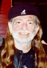 Head shot of Willie Nelson, with gray beard and long red hair and wearing a beret, smiles while looking at the camera.