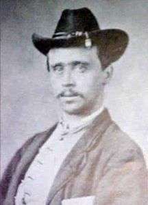 Head and shoulders of a white man with a thin mustache, wearing a cavalry hat, vest, and dark jacket.