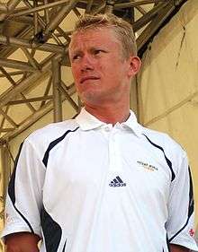 A blond-haired man in his mid-thirties, wearing a white polo shirt bearing an Adidas logo. He is looking to his right.