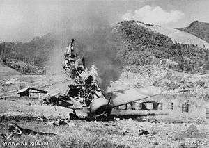 Propeller aircraft, nose pointing down, and ablaze. In the background are 44-gallon drums.