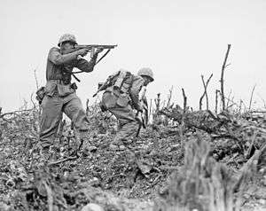 black & white photograph of two Marines advancing up a hill, the one on the left is firing an M1 submachinegun while the one on the right dashes for cover