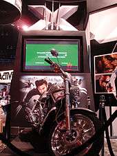 A silver motorcycle with black tires sits on a podium. A TV screen with a green preview displayed sits behind the motorcycle with a large X above it and a picture of Wolverine below