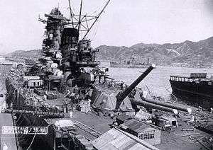 A view over a dock containing a large warship in the final stages of construction. Hills and a town can be seen across the harbor, a number of other ships are visible in the middle distance, and filling the foreground the warship's deck is littered with cables and equipment.
