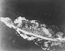 A close view of a large warship from almost directly overhead. Her wake is streaming out behind her and two trails of smoke are visible: a faint plume near her smokestack and a much thicker white plume partially obscuring her foremost main gun turret.