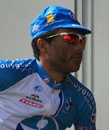 A black man of about thirty, wearing a blue and white cycling jersey with orange trim, a matching cap, and sunglasses.