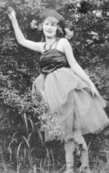 A black and white photograph of a young woman outdoors in a ballet pose, one arm extended. She looks at the camera.